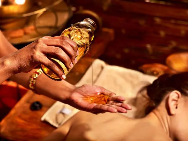 Рука массажиста, наливающего в ладонь масло на фоне лежащей девушки